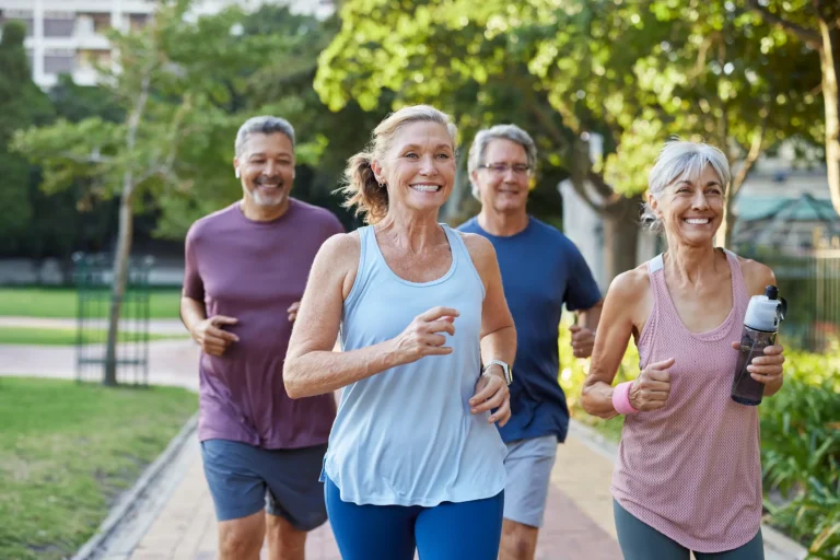 Four residents jogging together on a paved path in a park-like outdoor setting, enjoying a wellness activity.